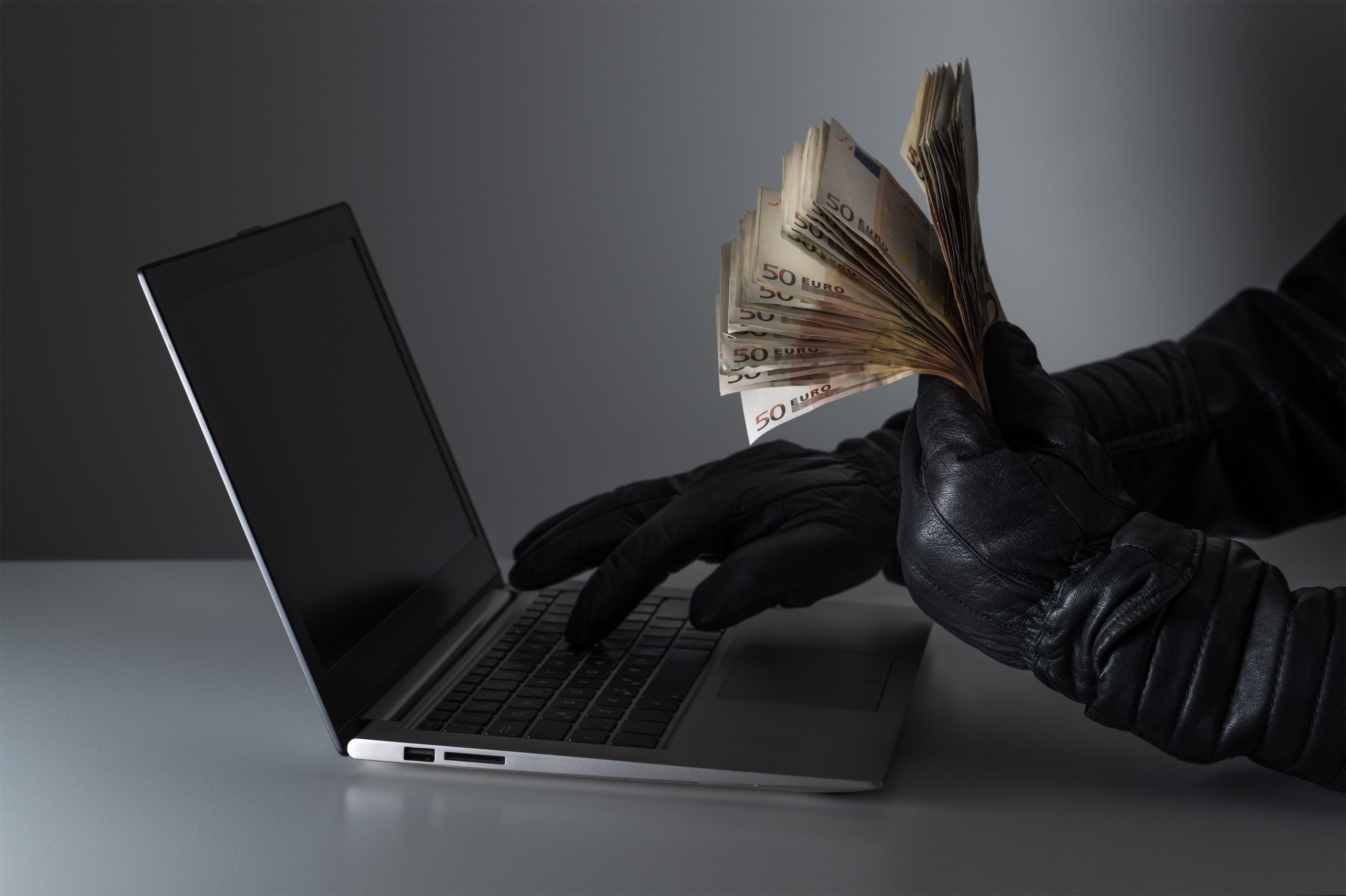 Internet fraud and cyber security concept. Criminal holding a lot of money and using laptop with black leather gloves.